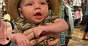 Theodore is registered to the contest to win money with this photo: baby, child, indoor, clothing_store, hat, straw_hat, camouflage, onesie, person, hands, face, curious, apparel, shopping, rack, wood_floor, display, retail, fashion, cute