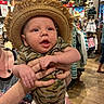 baby, child, indoor, clothing_store, hat, straw_hat, camouflage, onesie, person, hands, face, curious, apparel, shopping, rack, wood_floor, display, retail, fashion, cute