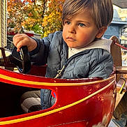 Jacob participe au concours pour gagner de l'argent avec cette photo : child, boy, blue_eyes, jacket, hoodie, amusement_ride, steering_wheel, red_car, autumn, fall_leaves, outdoor, daylight, portrait, cute, serious_expression, hair, seat, playground, fun, young