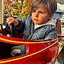 child, boy, blue_eyes, jacket, hoodie, amusement_ride, steering_wheel, red_car, autumn, fall_leaves, outdoor, daylight, portrait, cute, serious_expression, hair, seat, playground, fun, young