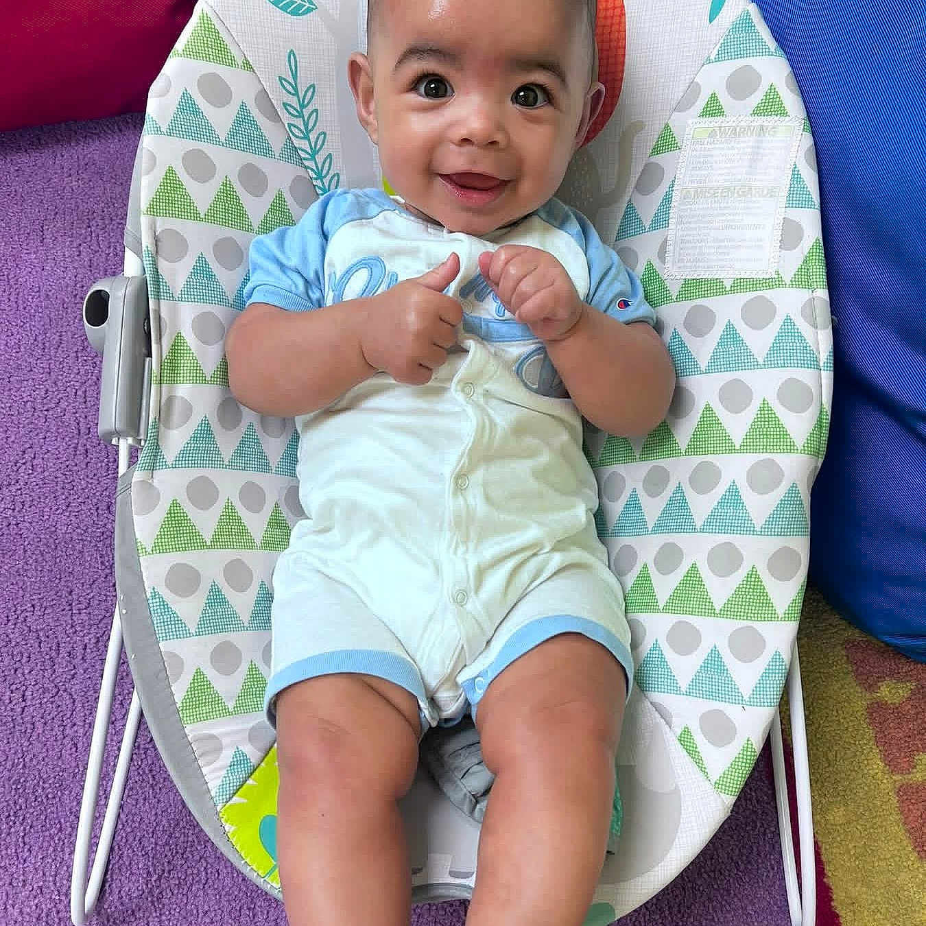 Zayden joined the competition — help win amazing prizes! baby, baby_rocker, barefoot, blue_clothing, child, colorful_background, cute, fabric_pattern, happy, head, indoor, infant, legs, person, playroom, purple_rug, seat, smiling, thumbs_up, young_child