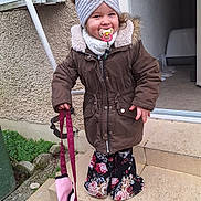 Irina participe au concours pour gagner de l'argent avec cette photo : toddler, child, coat, headband, pacifier, bag, steps, outdoor, smile, flowered_pants, doorway, concrete, wall, greenery, mailbox, winter_clothing, fur_hood, happy, person, daylight