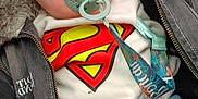 Clark participe au concours pour gagner de l'argent avec cette photo : baby, pacifier, superman_logo, jacket, blue_pants, infant, child, cute, clothing, seat, fabric, face, eyes, casual_wear, indoor, portrait, young_child, comfort, relaxing, superhero