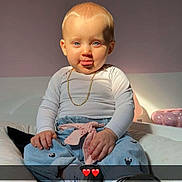 Sienna a rejoint le concours — aidez-le/la à gagner de superbes lots ! baby, child, tongue_out, blue_eyes, white_shirt, blue_pants, pink_bow, gold_chain, sitting, cushion, indoor, cute, playful, sunlight, portrait, person, footwear, smile, expression, heart_emoji