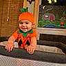 baby, child, pumpkin_costume, smiling, playpen, indoor, wooden_blinds, wall_decoration, painting, night_sky, campfire, camper, green_leaves, orange_clothing, happy, person, portrait, cute, holiday_costume, festive