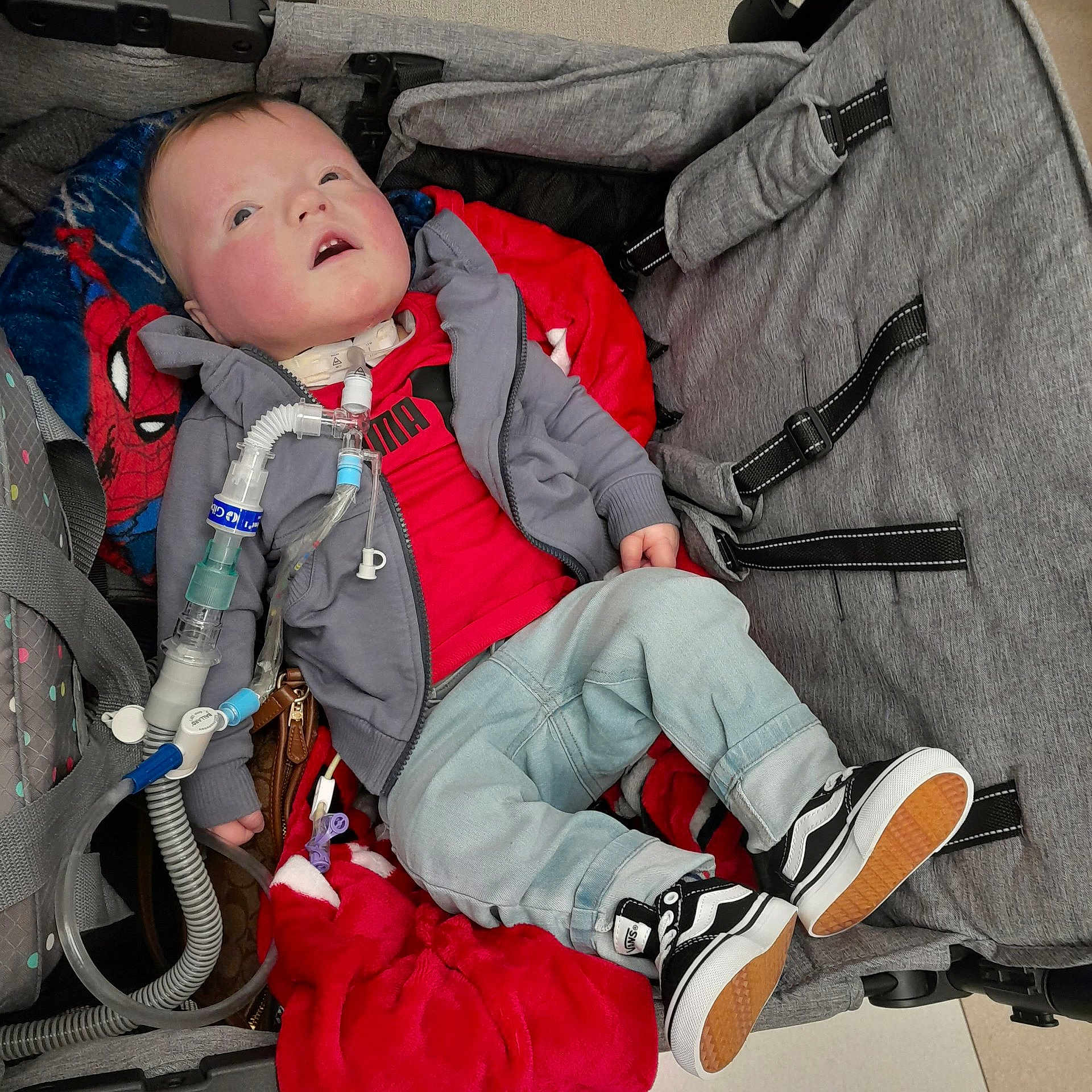 Bryant is registered to the contest to win money with this photo: child, baby, stroller, medical_equipment, blanket, spiderman, red_shirt, jeans, sneakers, black_and_white_shoes, indoor, resting, casual_clothing, medical_care, young_child, comfort, person, face, head, cozy