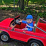 Isabella is registered to the contest to win money with this photo: toddler, child, toy_car, red_car, blue_clothing, blue_cap, outdoor, grass, dirt, fence, trees, sunlight, play, steering_wheel, backyard, small_shed, curious, vehicle, person, nature