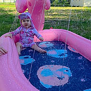 Isabella is registered to the contest to win money with this photo: toddler, child, swimsuit, inflatable_pool, pink, water_spray, dolphin_shape, outdoor, grass, trees, sunny, backyard, summer, play, water, fun, person, baby, sitting, pool