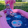 toddler, child, swimsuit, inflatable_pool, pink, water_spray, dolphin_shape, outdoor, grass, trees, sunny, backyard, summer, play, water, fun, person, baby, sitting, pool