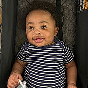 Dawson is registered to the contest to win money with this photo: baby, child, smiling, curly_hair, striped_clothing, onesie, pacifier, stroller, cushion, infant, happy, person, indoor, cute, portrait, young, hand, face, playful, comfort