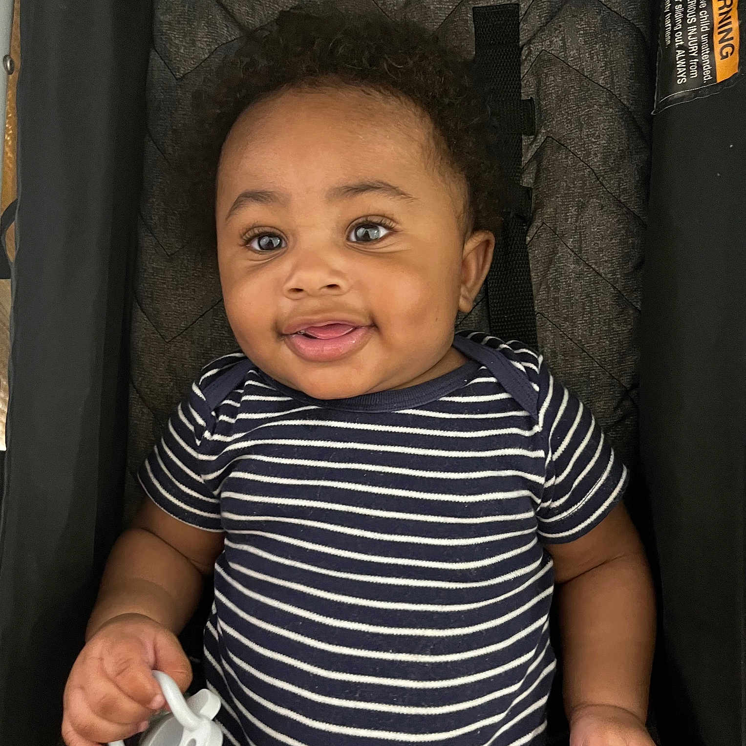 Dawson is registered to the contest to win money with this photo: baby, child, smiling, curly_hair, striped_clothing, onesie, pacifier, stroller, cushion, infant, happy, person, indoor, cute, portrait, young, hand, face, playful, comfort