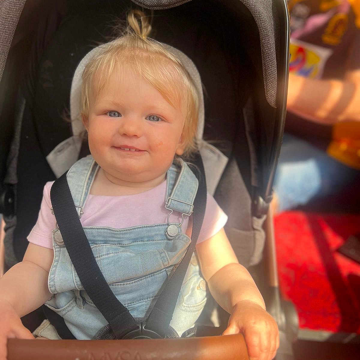 Harper is registered to the contest to win money with this photo: baby, blonde_hair, casual_clothing, child, denim_overalls, grip, handlebar, happy, indoor, person, pink_shirt, portrait, safety_harness, seat, seatbelt, smiling, stroller, sunlight, toddler, young_child