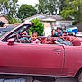 child, toddler, car, convertible, vintage_car, red_car, braided_hair, outdoor, suburban, vehicle, street, tree, house, playful, person, side_view, summer, transport, curious, parked_car