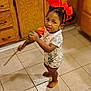 toddler, child, flower, red_bow, hair_accessory, barefoot, kitchen, tile_floor, wooden_cabinets, curious, standing, onesie, indoor, person, young_child, orange_flower, cute, home, portrait, baby