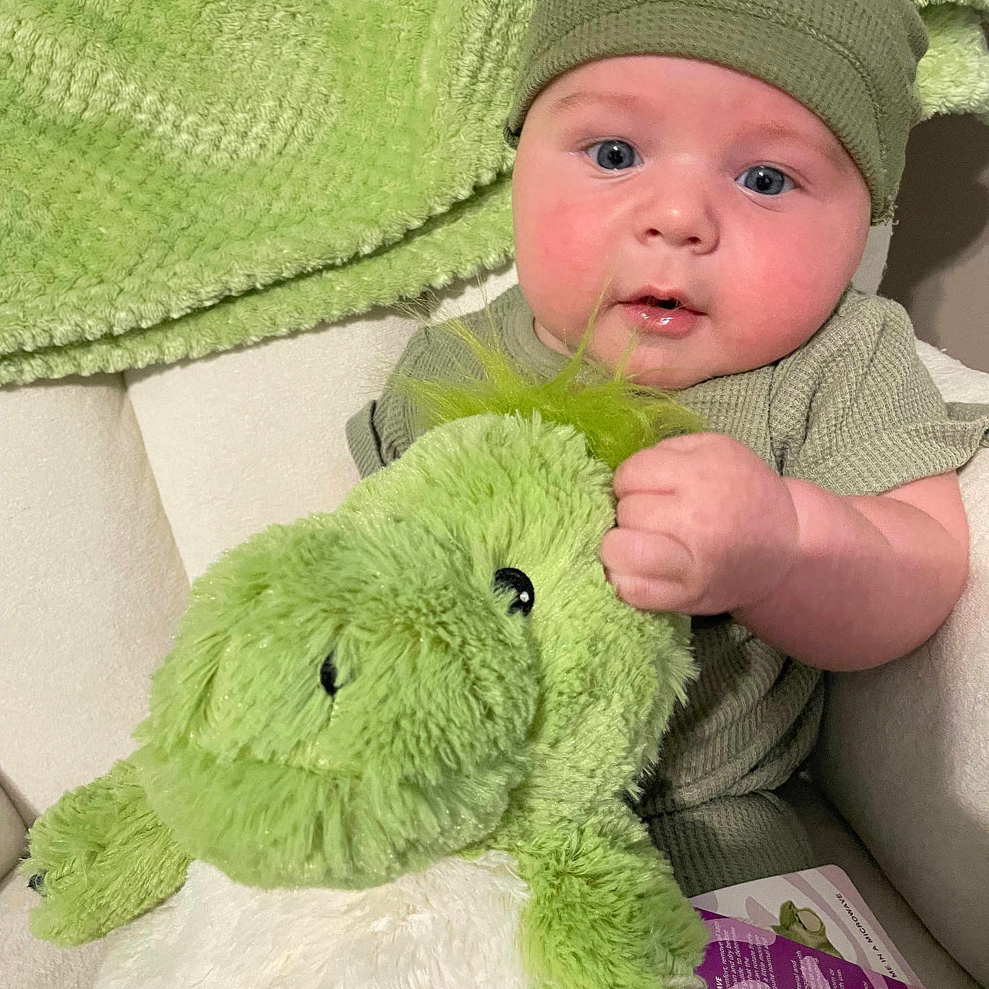 Wylde is registered to the contest to win money with this photo: baby, child, plush_toy, frog, green_clothing, hat, blanket, soft_texture, cute, indoor, portrait, infant, holding, stuffed_animal, cozy, closeup, blue_eyes, chubby_cheeks, seated, curious