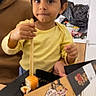child, toddler, yellow_shirt, chopsticks, sushi, food, box, table, kitchen, indoor, person, face, brown_hair, expression, hand, seat, curious, eating, casual, home