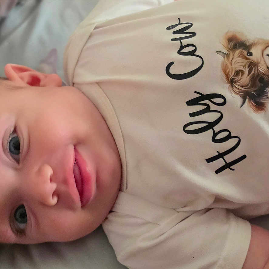 Airis Elaine is registered to the contest to win money with this photo: animal_illustration, baby, bed, blanket, calf, child, clothing, cute, expression, face, happy, head, highland_cow, indoors, infant, lying_down, person, skin, smiling, tshirt