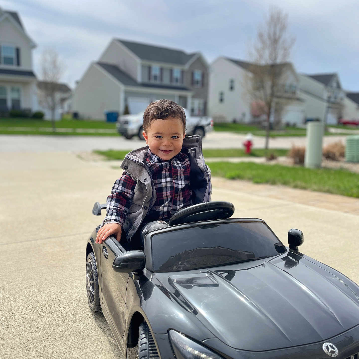 Amar is registered to the contest to win money with this photo: black_car, car_toy, child, curly_hair, daytime, driveway, green_grass, happy, house, outdoor, person, plaid_shirt, playing, sidewalk, smiling, suburban, toddler, toy_car, vehicle, vest