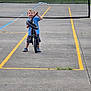 child, bike, outdoor, concrete, tennis_net, grass, court, smiling, blue_shirt, shorts, sneakers, playful, fence, daylight, young, person, recreation, sport, standing, fun