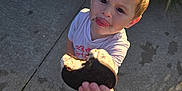 Thomas is registered to the contest to win money with this photo: child, toddler, cookie, oreo, food, hand, face, tongue, crumbs, outdoor, sidewalk, grass, plant, sunlight, casual_clothing, playful, offering, person, young_child, footwear