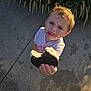 child, toddler, cookie, oreo, food, hand, face, tongue, crumbs, outdoor, sidewalk, grass, plant, sunlight, casual_clothing, playful, offering, person, young_child, footwear