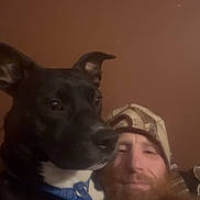 Kali joined the competition — help win amazing prizes! dog, black_dog, human, man, beard, cap, selfie, portrait, indoor, low_light, close_up, pet, blue_collar, brown_background, companionship, sitting, face, animal, casual, photograph