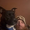 dog, black_dog, human, man, beard, cap, selfie, portrait, indoor, low_light, close_up, pet, blue_collar, brown_background, companionship, sitting, face, animal, casual, photograph