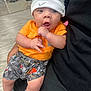 Ramontez joined the competition — help win amazing prizes! baby, infant, beanie, orange_shirt, patterned_shorts, gray_socks, hand_gesture, indoor, person_holding, flooring, surprised_expression, headwear, clothing, cute, child, portrait, young_child, seated, casual, fabric