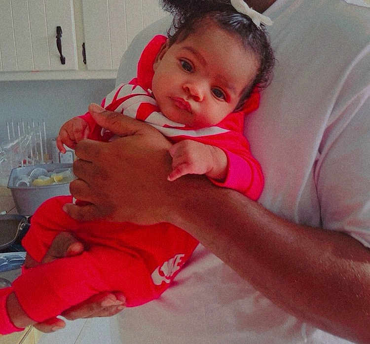 Marjae is registered to the contest to win money with this photo: baby, infant, red_clothing, nike, person, adult_hand, kitchen, cabinet, countertop, dishrack, curly_hair, casual, indoor, portrait, holding, calm, looking_at_camera, white_shirt, skin_tone, close_up