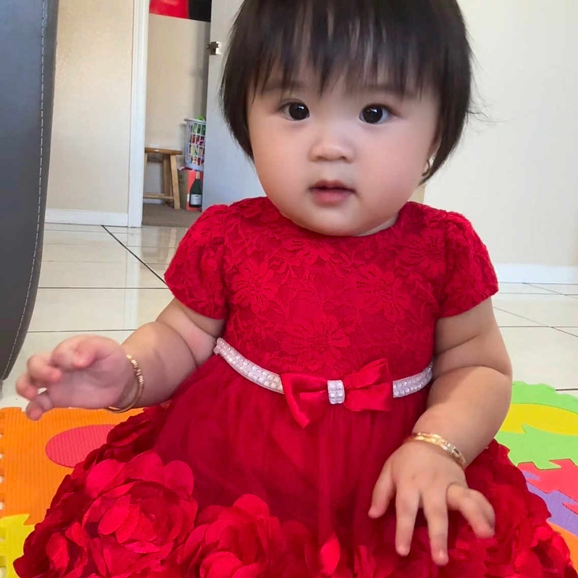 Mimi Nguyen is registered to the contest to win money with this photo: baby, black_hair, bracelet, child, colorful, curious, cute, door, floor_tiles, flower_clip, home_interior, indoor, person, pink, play_mat, portrait, red_dress, sitting, wall_poster, young_child