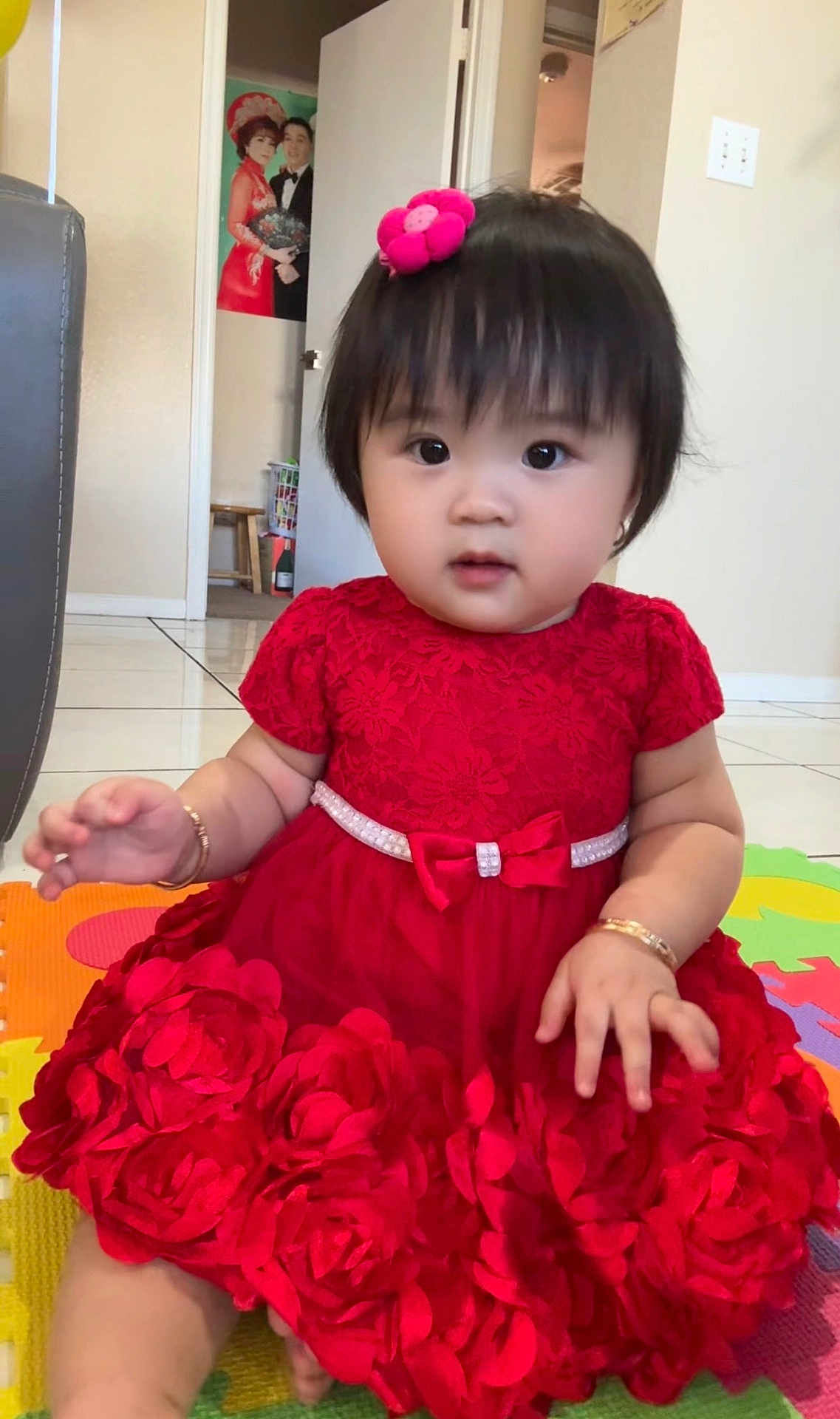 Mimi Nguyen is registered to the contest to win money with this photo: baby, child, red_dress, flower_clip, play_mat, indoor, portrait, cute, sitting, curious, home_interior, floor_tiles, bracelet, pink, black_hair, young_child, wall_poster, door, colorful, person