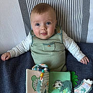 Blandine a rejoint le concours — aidez-le/la à gagner de superbes lots ! baby, child, blue_eyes, green_outfit, fabric_book, soft_toy, striped_pillow, cute, smiling, indoor, infant, sitting, playful, young_child, person, portrait, hands, cheeks, clothing, toy