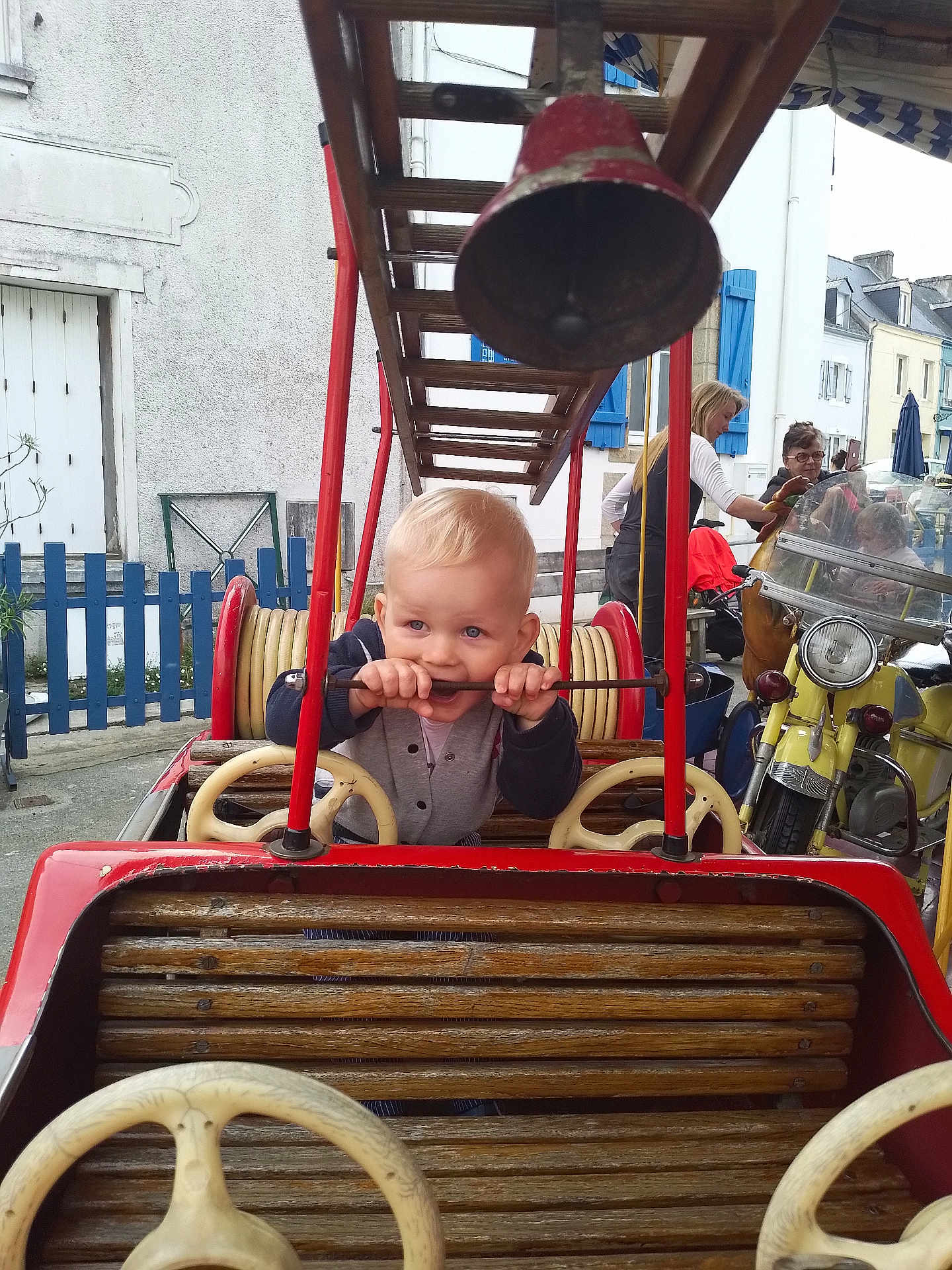 Aodren participe au concours pour gagner de l'argent avec cette photo : adult, blonde_hair, building, child, curious, daylight, fence, fire_truck, helmet, motorcycle, outdoor, people, playground, red, steering_wheel, toddler, toy_car, vintage, window, wood
