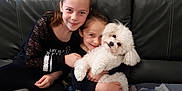 Naya participe au concours pour gagner de l'argent avec cette photo : child, dog, indoor, smiling, couch, hugging, pet, white_dog, casual_clothing, plant, wooden_door, family, happy, leather_couch, home, sitting, portrait, two_children, cozy, friendly