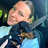 woman, dog, puppy, dachshund, car, seatbelt, white_shirt, smile, portrait, indoor, pet, holding, black_dog, tan_markings, human_hand, short_hair, seat, window, daylight, closeup