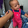 toddler, child, overalls, red_stripes, blue_shirt, smiling, hand, bracelet, bed, blanket, indoor, furniture, person, happy, cute, curly_hair, skin, room, floor, casual