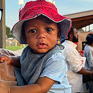 Nasir Cayden is registered to the contest to win money with this photo: baby, child, bucket_hat, red_hat, spiderman, blue_clothing, person, face, eyes, outdoor, daylight, people, background, holding, portrait, curious, expression, hand, shoulder, hat