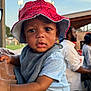 baby, child, bucket_hat, red_hat, spiderman, blue_clothing, person, face, eyes, outdoor, daylight, people, background, holding, portrait, curious, expression, hand, shoulder, hat