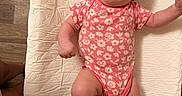 Gabrielle is registered to the contest to win money with this photo: baby, child, infant, smiling, portrait, flower_crown, pink_outfit, floral_pattern, onesie, socks, heart_design, lying_down, changing_pad, quilted_surface, indoor, wood_floor, adult_partial, cheerful, baby_fashion, playful