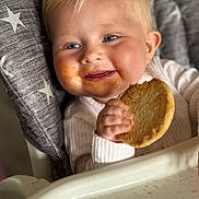 Lillian is registered to the contest to win money with this photo: baby, blonde_hair, blue_eyes, child, cookie, crumbs, cute, eating, face, hand, happy, high_chair, indoor, infant, messy_face, portrait, smiling, snack_time, star_pattern, white_clothing