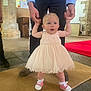 adult_hands, baby, black_pants, blue_shirt, carpet, casual_shoes, child, church, cute, dress, floor, helping, indoor, person, pink_shoes, socks, stone_wall, support, toddler, walking