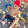 child, toddler, beach, sand, toy, red_cap, shorts, barefoot, sunlight, shadow, chair, towel, sand_toys, playing, summer, outdoor, vacation, footwear, plastic_toy, relaxation