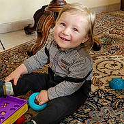 Elyo participe au concours pour gagner de l'argent avec cette photo : toddler, child, smiling, indoor, toy, plaid_shirt, dark_pants, blue_ring, carpet, patterned_rug, wooden_furniture, floor, playful, happy, sitting, footwear, cute, person, home, bright_eyes