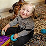 toddler, child, smiling, indoor, toy, plaid_shirt, dark_pants, blue_ring, carpet, patterned_rug, wooden_furniture, floor, playful, happy, sitting, footwear, cute, person, home, bright_eyes
