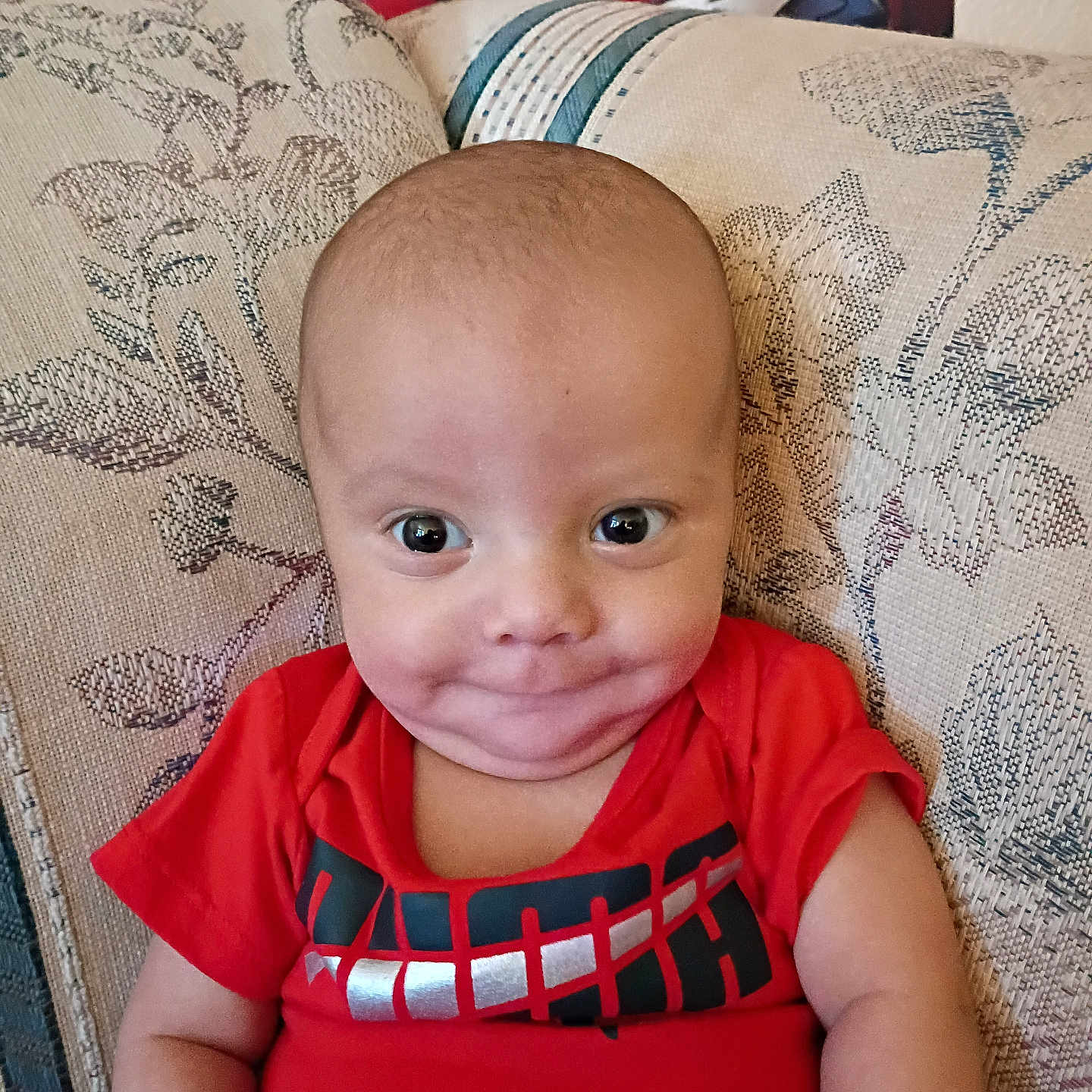 Mason is registered to the contest to win money with this photo: baby, child, couch, cute, expression, eyes, face, furniture, happy, head, indoor, patterned_fabric, person, portrait, red_shirt, room, sitting, skin, smile, young