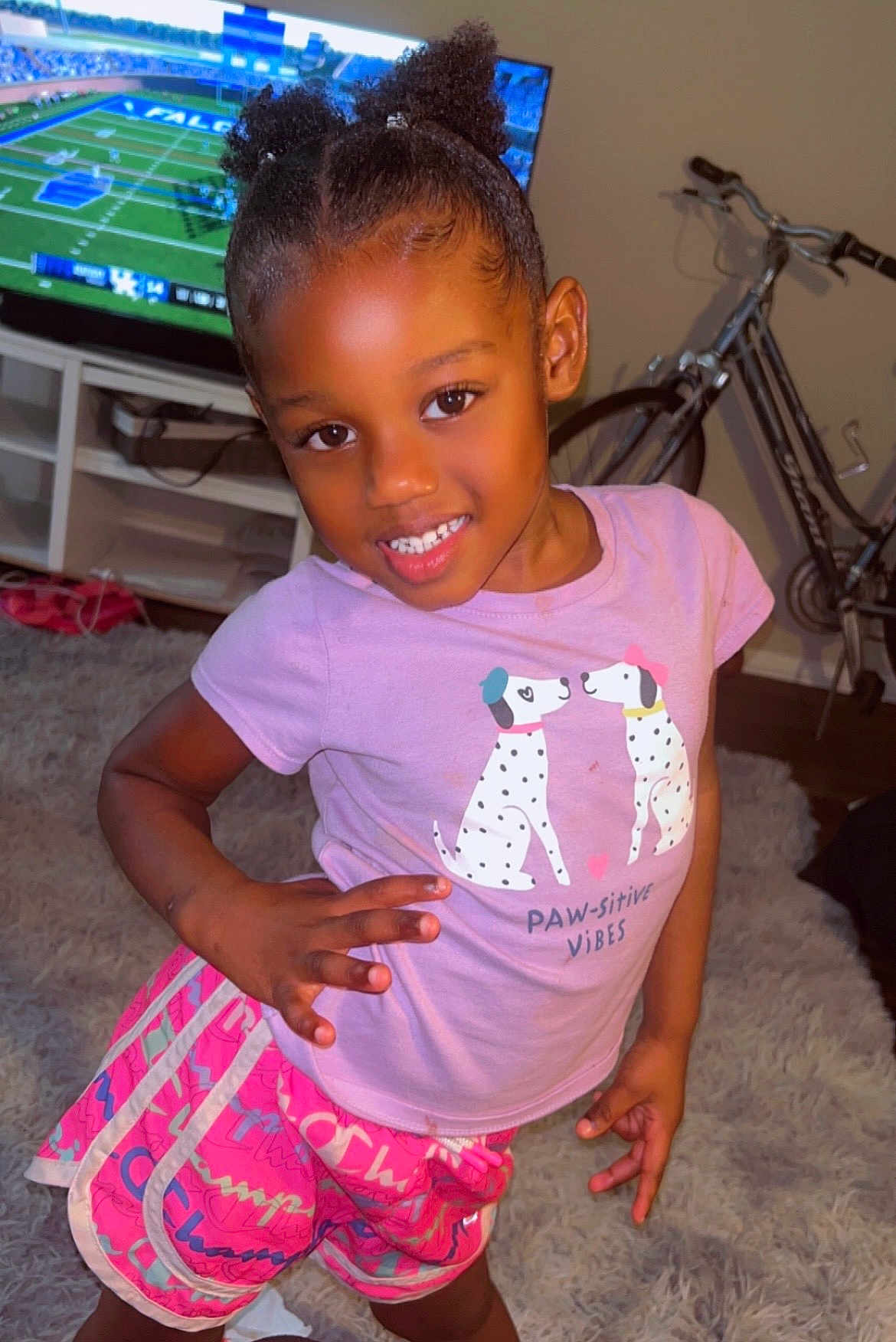 Lamyiah joined the competition — help win amazing prizes! child, girl, smiling, indoor, pink_shirt, shorts, dog_print, television, football_game, bicycle, carpet, hand_on_hip, happy, person, hair_puffs, room, furniture, casual, pose, home