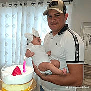 Danna Camila is registered to the contest to win money with this photo: man, baby, birthday_cake, candle, strawberry, white_clothing, headband, bow, indoor, curtains, smile, holding, celebration, portrait, family, person, wooden_floor, table, home, light