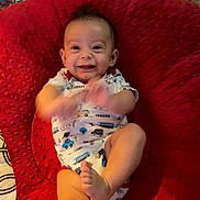 Zayden is registered to the contest to win money with this photo: baby, infant, smiling, clapping, cushion, red, onesie, patterned_clothing, foot, happy, indoor, child, cute, face, person, baby_girl, baby_boy, playful, soft, texture