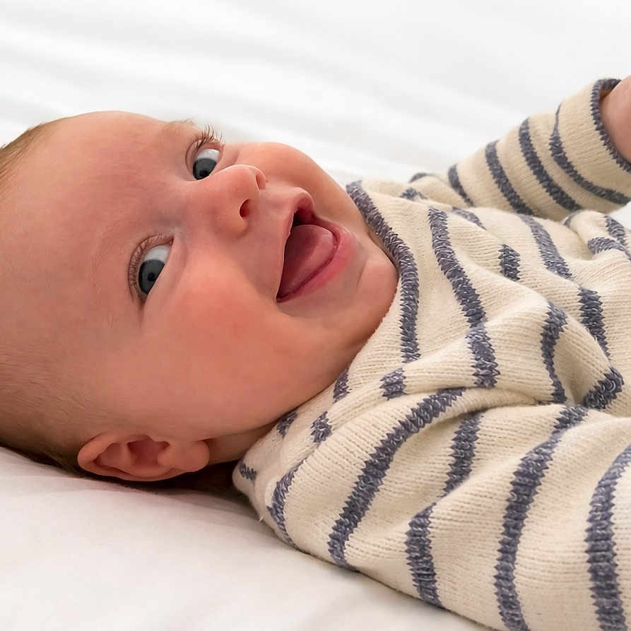 Oscar a rejoint le concours — aidez-le/la à gagner de superbes lots ! baby, smiling, striped_sweater, lying_down, white_bed, happy, child, infant, cute, face, person, cozy, eyes, mouth, skin, head, hand, portrait, indoors, baby_clothes