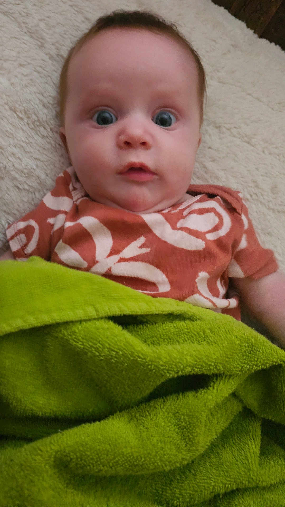 Zoie joined the competition — help win amazing prizes! baby, infant, child, blanket, green_blanket, orange_shirt, patterned_clothing, wide_eyes, face, head, lying_down, soft_surface, expression, indoor, person, cute, young_child, skin, portrait, comfort